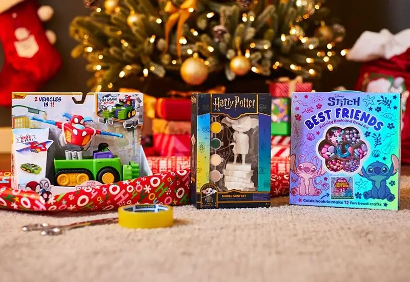 Three boxed craft and toy sets are displayed on a carpet in front of a decorated Christmas tree with wrapped presents. The items include a vehicle toy set with two cars, a Harry Potter model paint set, and a Stitch-themed “Best Friends” craft kit. Wrapping paper, tape, and a pen are also visible in the foreground.